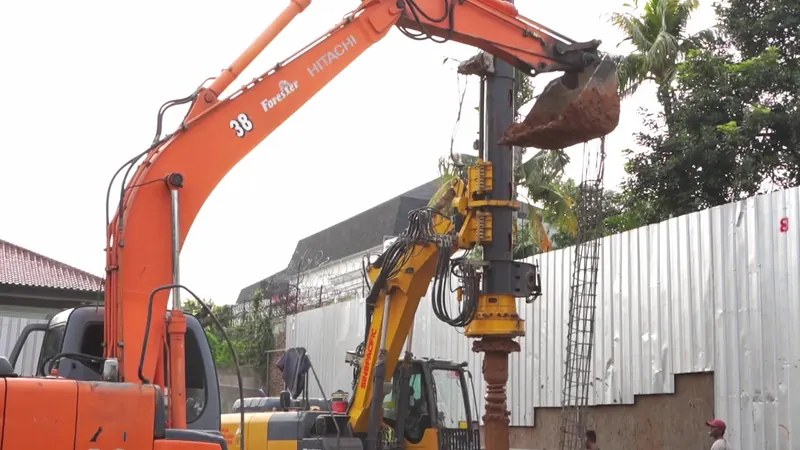 Rig bor dan excavator bekerja berdampingan di lahan sempit di balik pagar seng